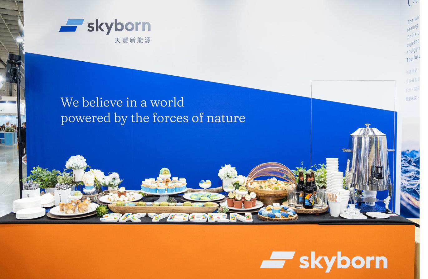 Skyborn 展位前的甜點布置：藍色品牌牆「We believe in a world powered by the forces of nature」之下，是白色甜點、藍綠色海洋餅乾、橘色 Skyborn 字母蛋糕，與品牌色精準呼應。