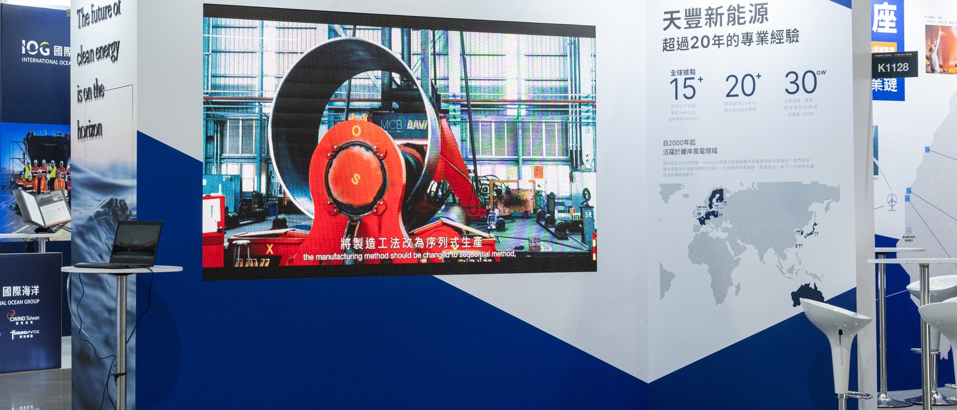 Skyborn 展位的水平全景：左側「The future of clean energy is on the horizon」直書文字牆、中央巨幅製造工法影片、右側天豐新能源資訊牆與全球地圖。三個展示平面在小空間裡折疊成 360 度的視野，每盞燈、每一吋木板的尺度都被精算。