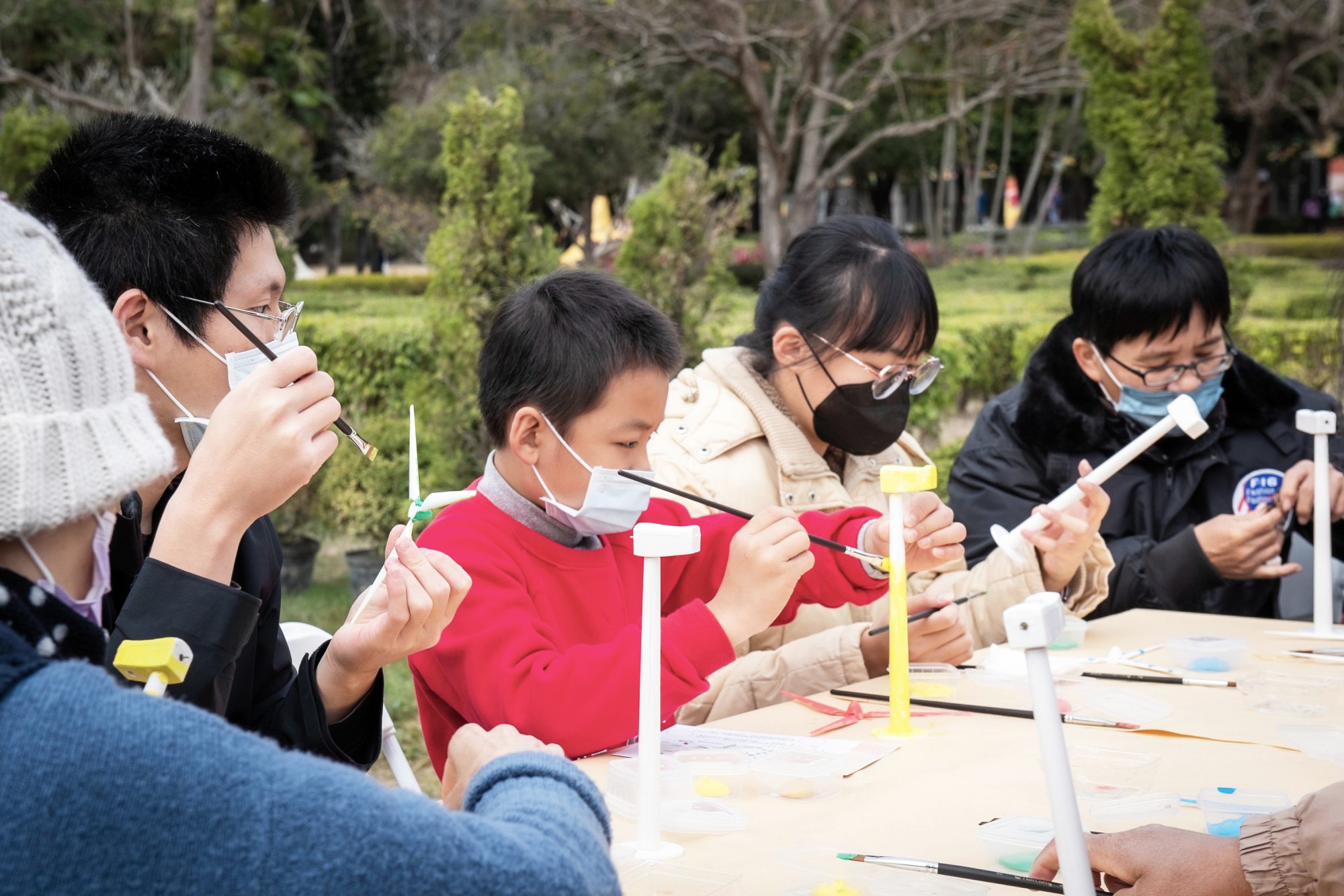 四個人並排坐在長桌前，各自專注地用畫筆為手中的白色微型風機塗上顏色。