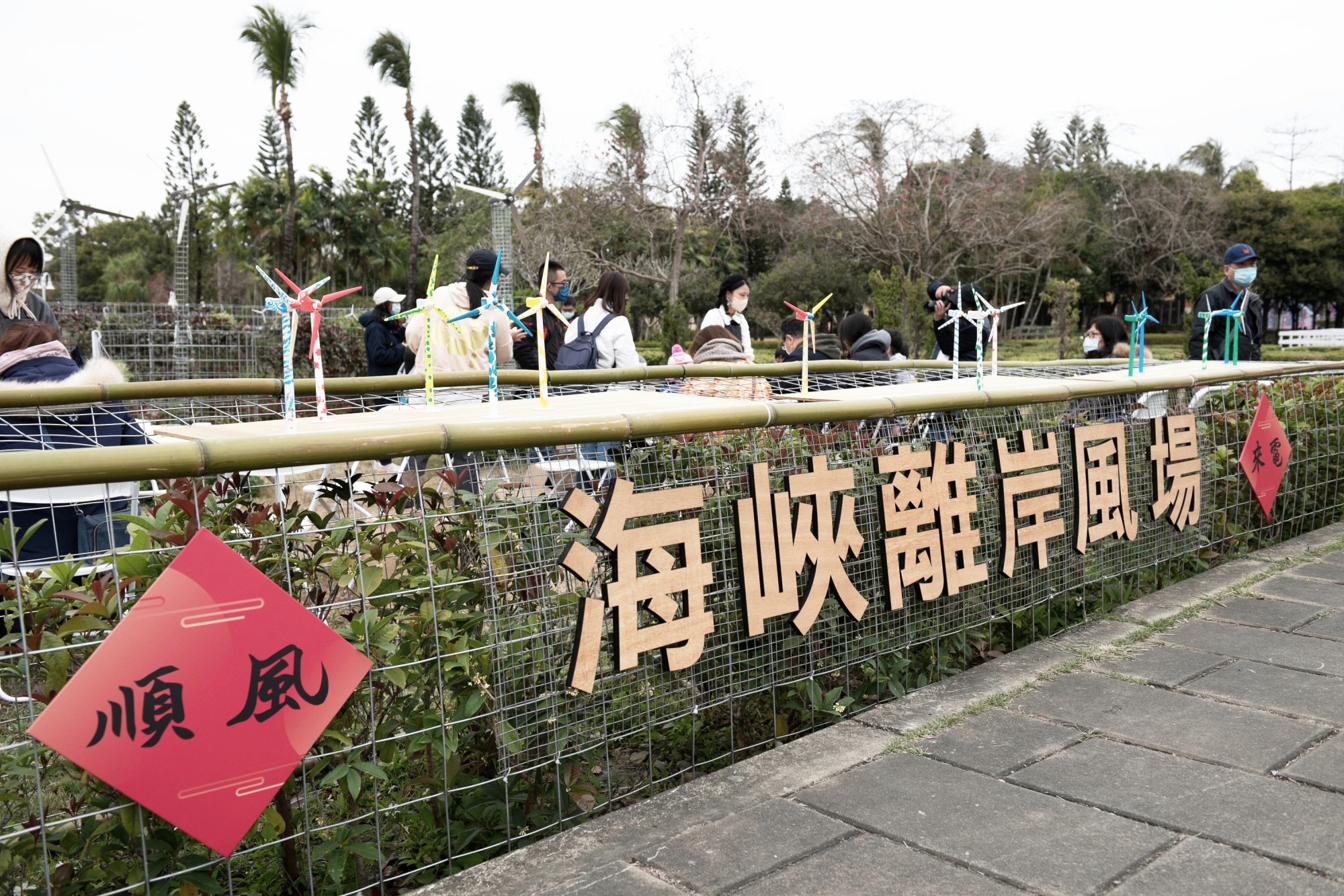 一排彩繪完成的小風機立在竹製平台上，後方木板字「海峽離岸風場」與紅色「順風」春聯——「順風來電」的物件成果。