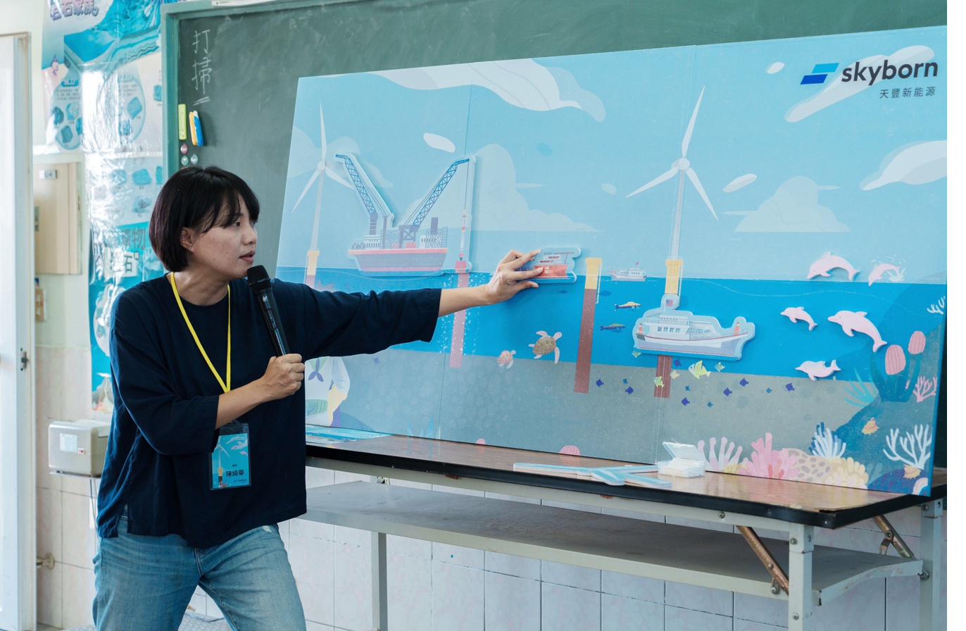 講師站在大型磁吸教學牆前，正將離岸風電工作船的磁吸件貼到牆面海平面位置——牆上是離岸風機、白海豚、海洋生態的插畫。