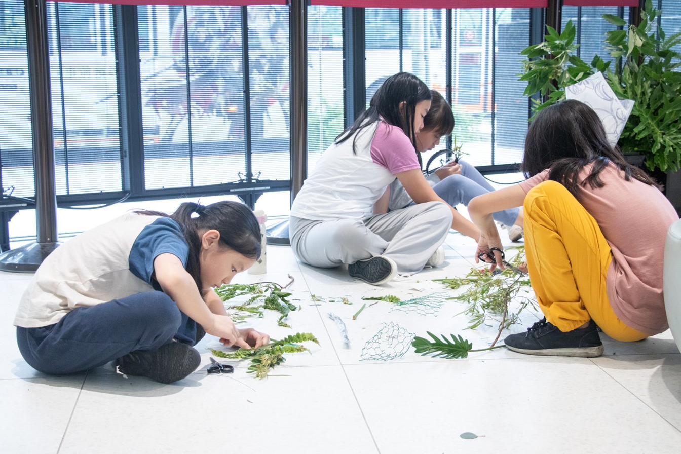 三位孩童席地而坐，正在裁剪與排列植物素材。地上散落著蕨葉、藍色網狀枝條與綠色葉子──每一片配飾，都是自己親手挑選與構築的。
