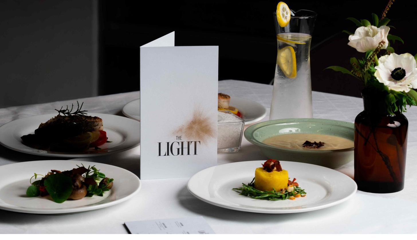 THE LIGHT 餐廳的桌面：白桌布、四盤精心擺盤的菜餚、立著的菜單、檸檬水壺、白色花藝。暗背景襯托出柔光與餐廳氛圍。