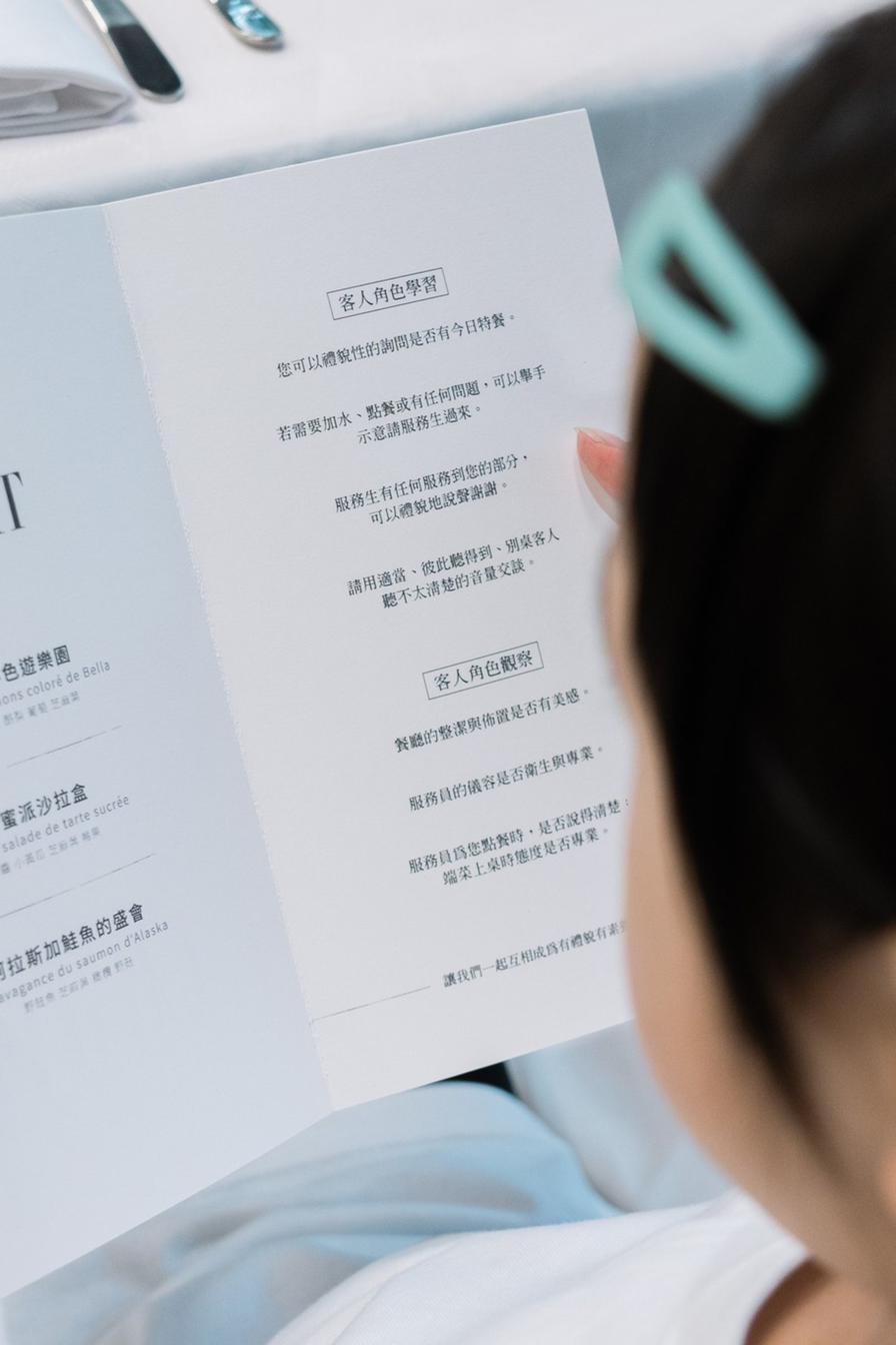 一位戴著藍色髮夾的孩童正在翻閱 THE LIGHT 菜單，紙上印有「客人角色學習」「客人角色觀察」等專業評鑑準則。